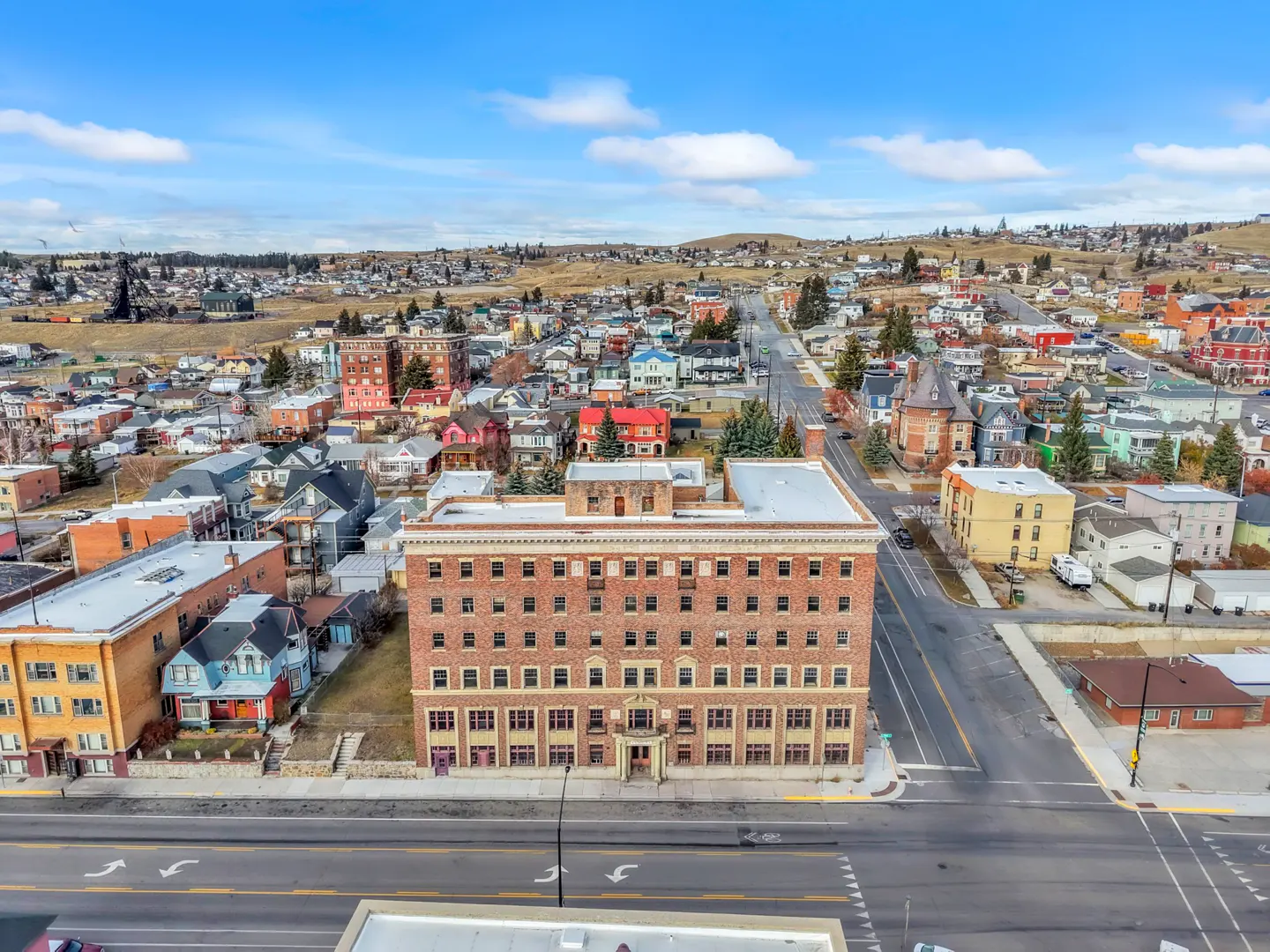 Aerial view of a four-story brick building in a town, with colorful houses and rolling hills in the background under a blue sky.