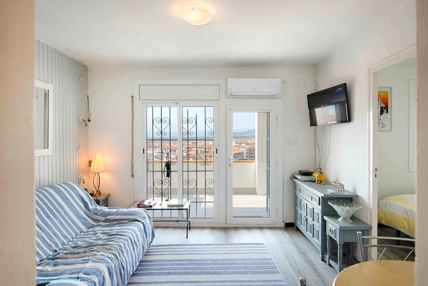Bright living room with a blue-striped sofa, matching rug, grey wooden furniture, and a balcony door with city views.