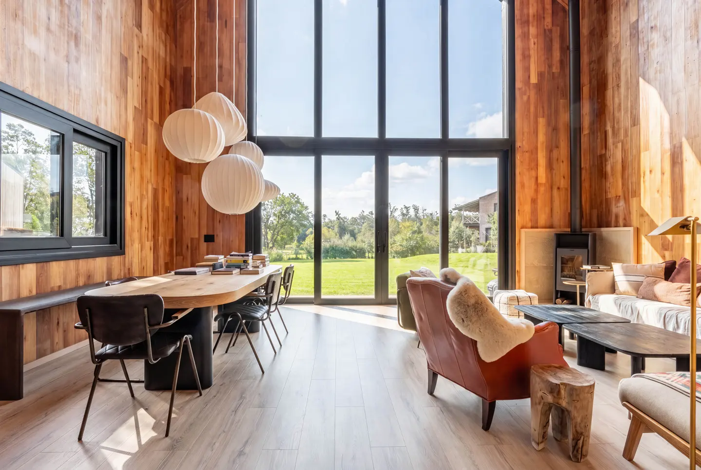 Modern living room with wood-paneled walls, floor-to-ceiling windows, a dining table, and cozy seating overlooking a lawn.