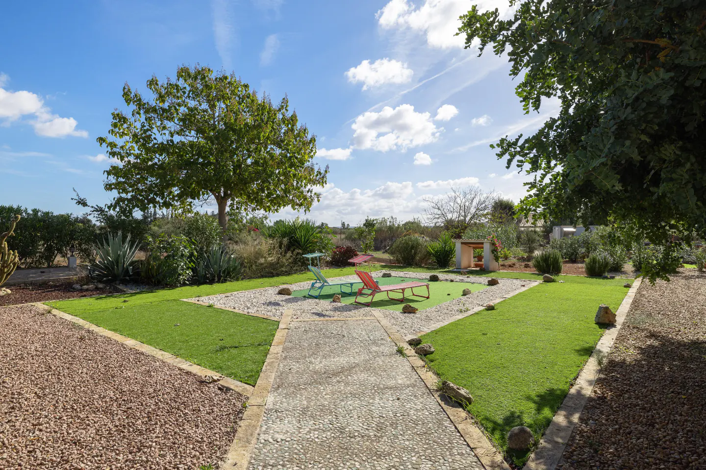 A sunny backyard with green grass, gravel, and a stone path. Two lounge chairs sit on a green patch surrounded by white gravel. A tree and blue sky are in the background.