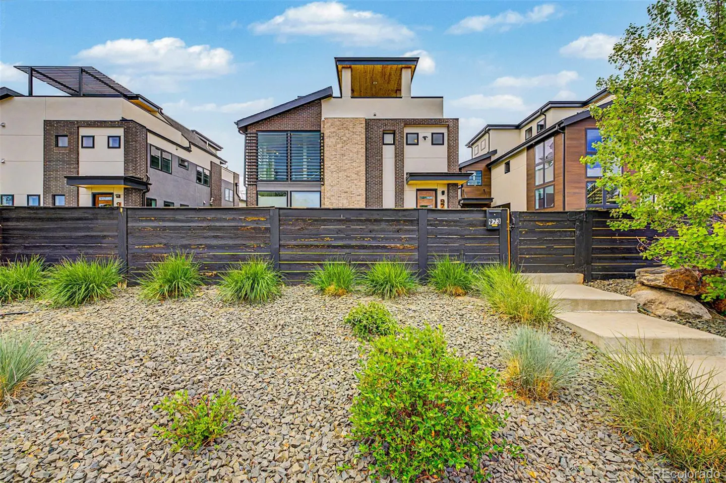 Modern two-story home with brick and beige siding, a dark wood fence, and a rock garden with green plants. Blue sky above.