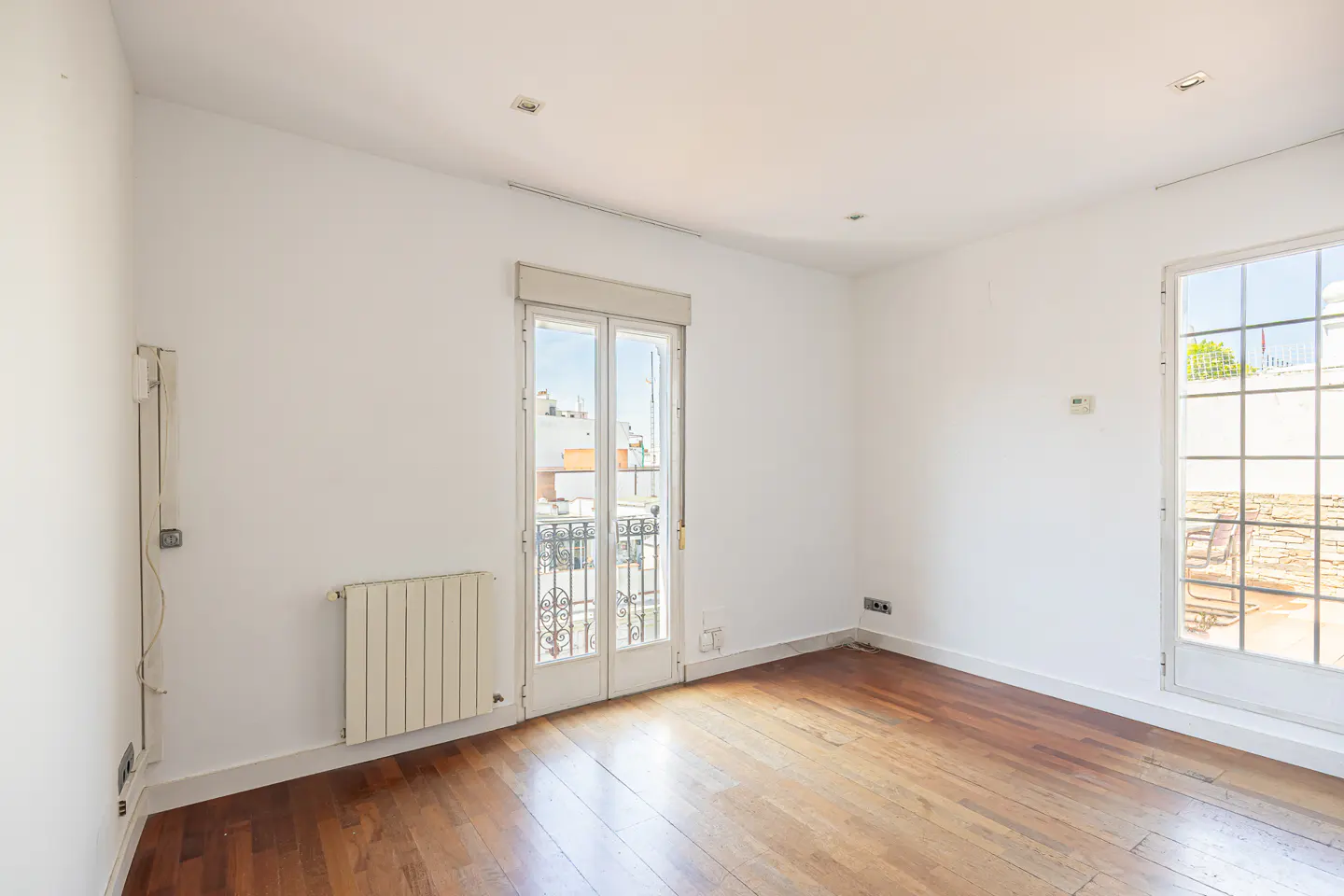 Bright, empty room with white walls, hardwood floors, and two white-framed windows. One window has a small balcony with black iron railings.
