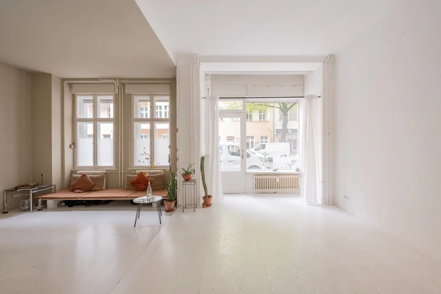 Bright, airy room with white walls and floors. Windows with frosted glass, a low platform bed, and plants add warmth. Street view visible.