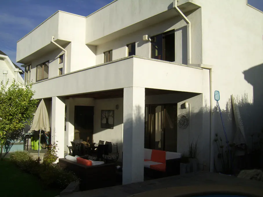 Exterior view of a modern, two-story white house with a covered patio, outdoor furniture, and a glimpse of a pool.