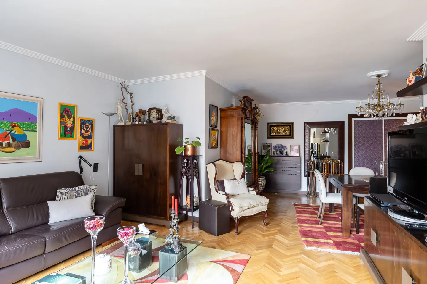 Living room with a brown leather sofa, cream armchair, wooden cabinets, dining area, and colorful art on light gray walls.