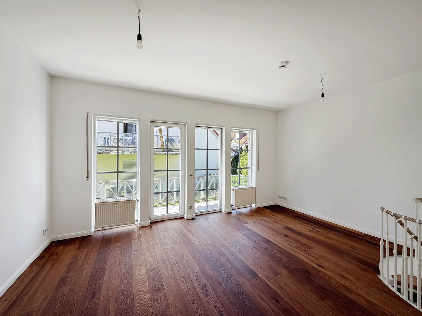 Bright, empty room with dark wood floors, white walls, and three windows with black frames. A white staircase is visible in the corner.