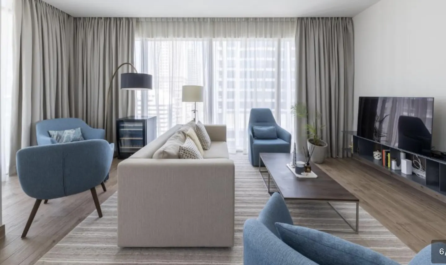 Living room with blue chairs, a beige sofa, and a dark wood coffee table on a striped rug. A TV sits on a shelf against a white wall.