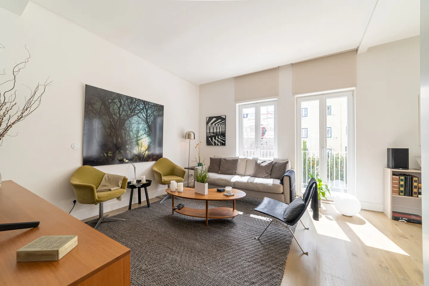 Bright living room with a gray rug, olive chairs, a gray sofa, and a wooden coffee table. Large windows let in sunlight.
