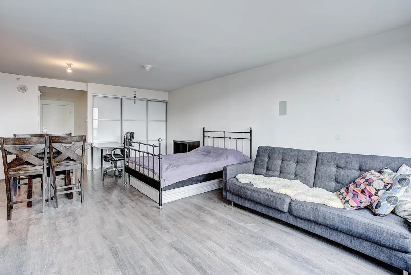 Studio apartment with a grey sofa, a bed with a black metal frame, a dining table with chairs, and light wood flooring.