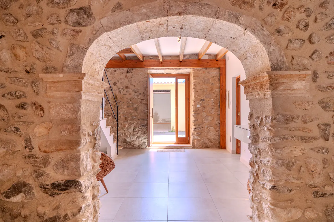 A stone archway frames a bright room with light tile floors, exposed wooden ceiling beams, and an open door to a patio.