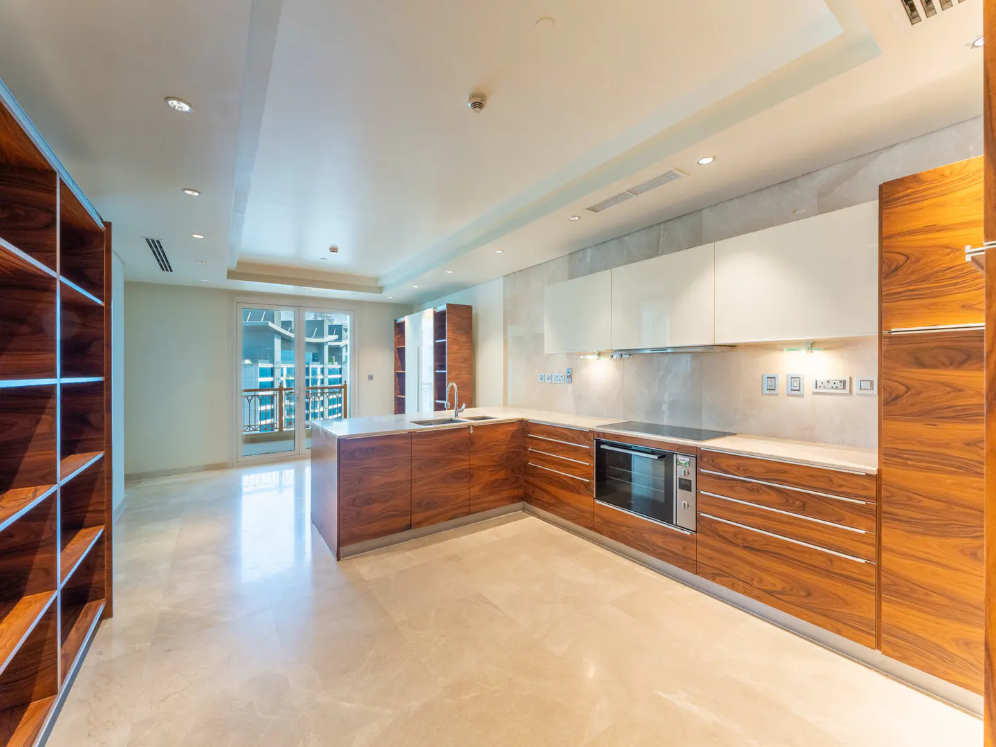 A modern kitchen with wood cabinets, white countertops, and marble floors. A balcony is visible through glass doors.