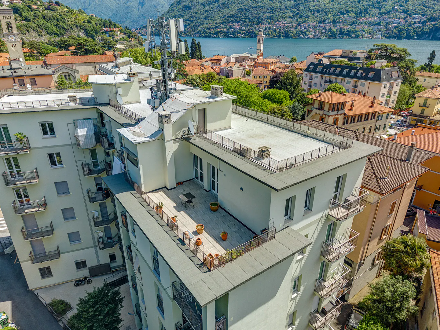 Aerial view of a white apartment building with a rooftop terrace, overlooking a town and lake with mountains in the background.