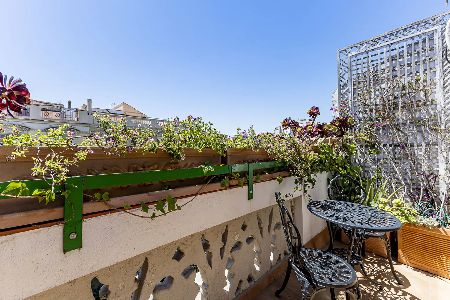 A sunny balcony with a black metal table and chairs, potted plants, and a white lattice screen.