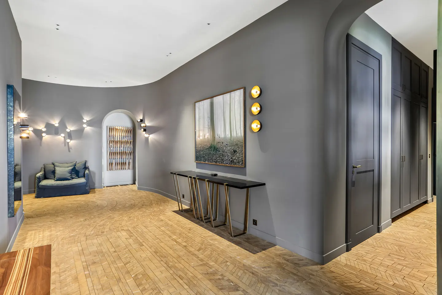 Modern hallway with grey curved walls, herringbone wood floors, a console table, forest art, and a cozy blue armchair.