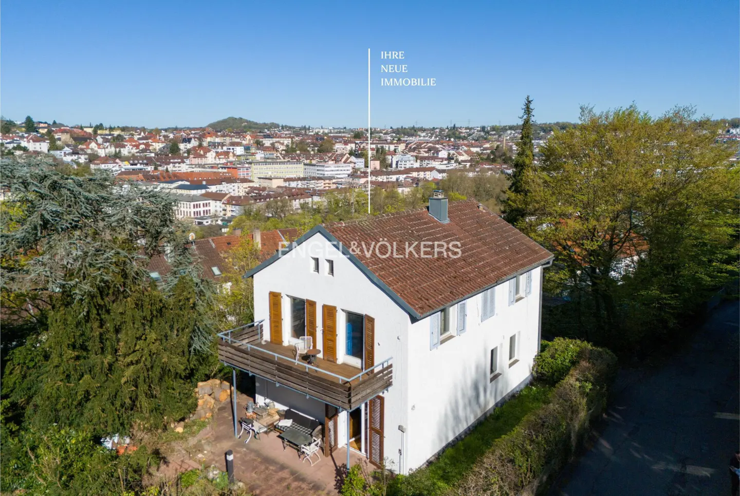 Two-story white house with a brown roof, balcony, and patio, surrounded by trees, with a city skyline in the background.