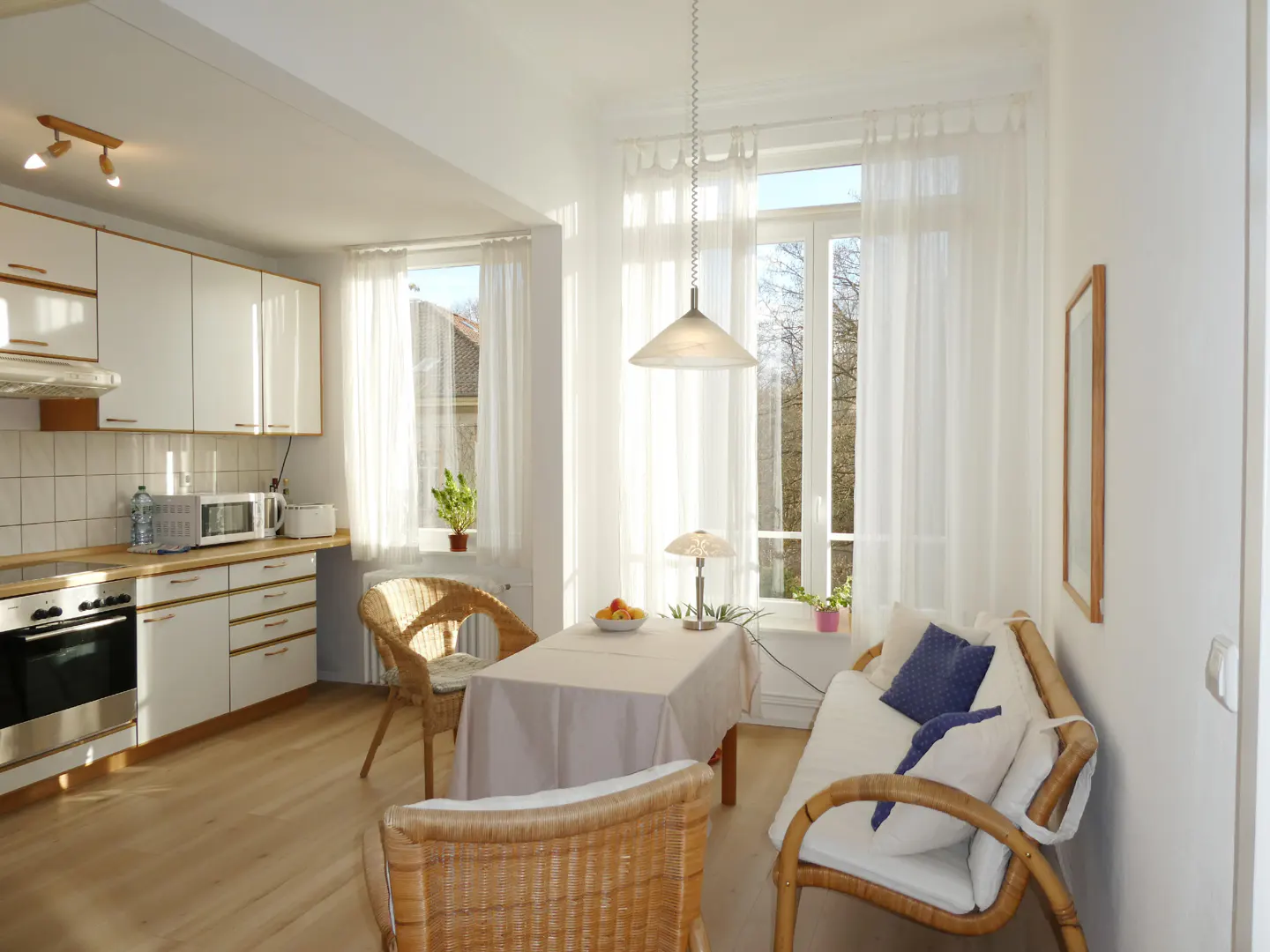 Bright, sunny kitchen and dining area with white cabinets, wicker chairs, and a table set for a meal. Large windows with sheer curtains.