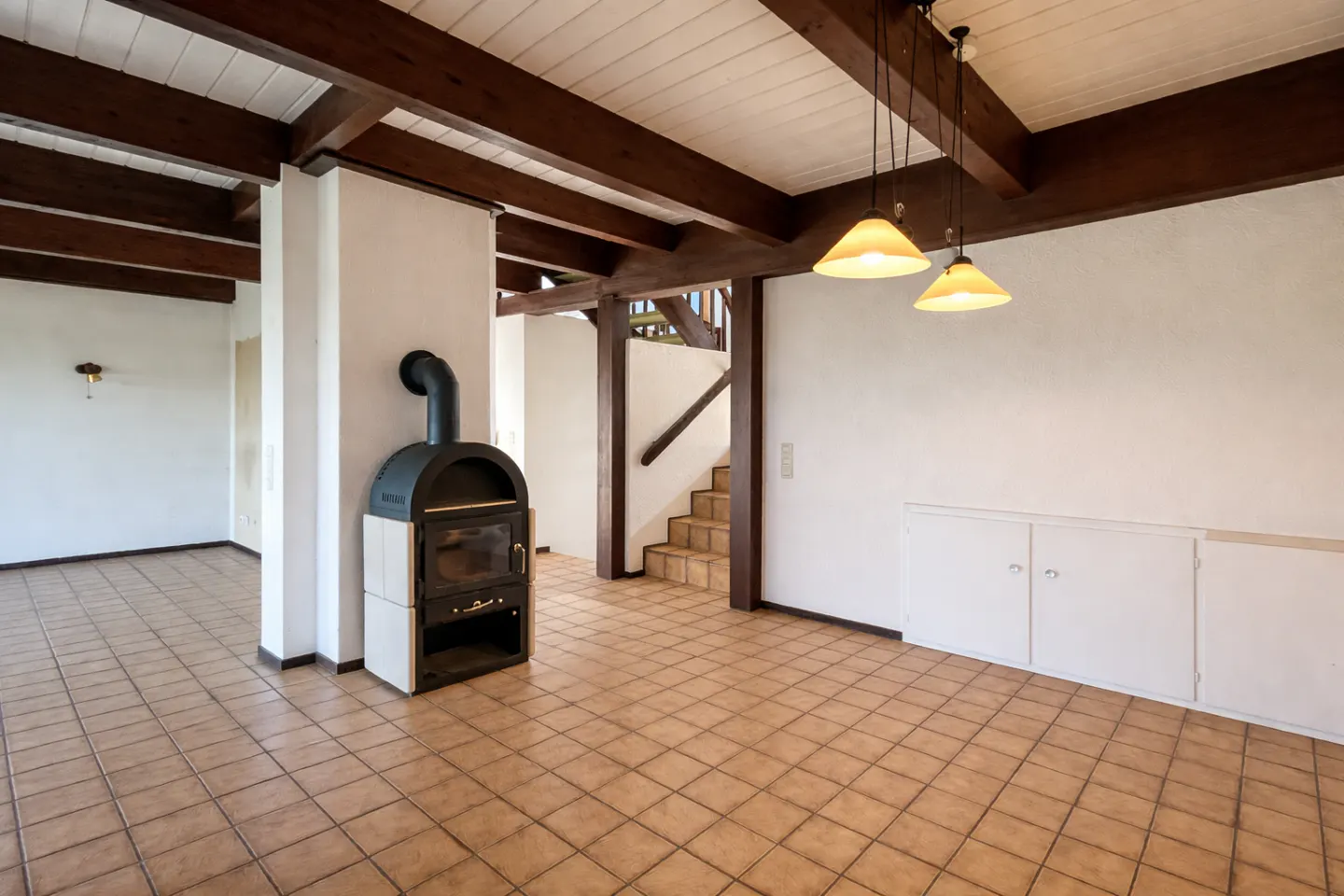A living room with a black fireplace, brown tile floor, wooden beams, and a staircase.
