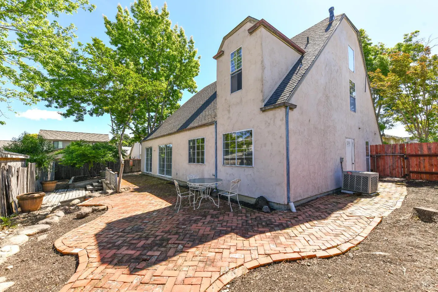 Backyard view of a two-story beige house with a brick patio, table, chairs, and green trees.