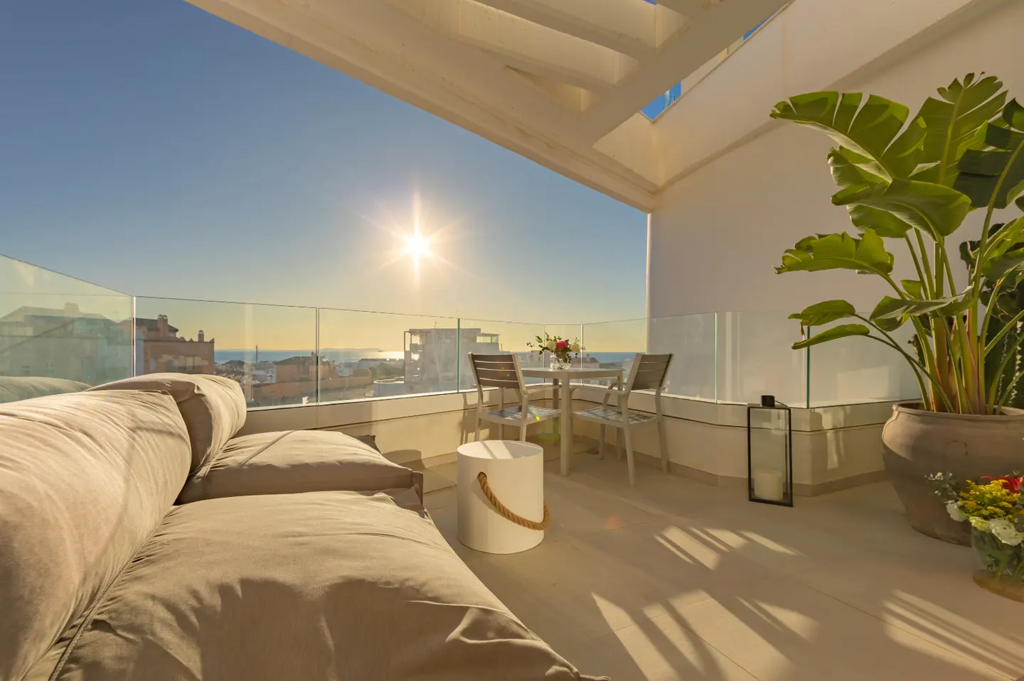 Sunny balcony with a beige sofa, table, chairs, and potted plants overlooking a city and the sea.