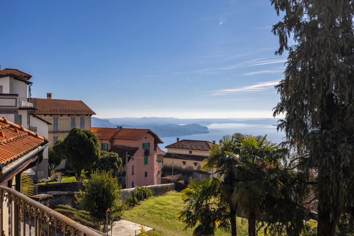 View of Italian hillside homes with red tile roofs, green grass, and a lake in the distance under a blue sky.