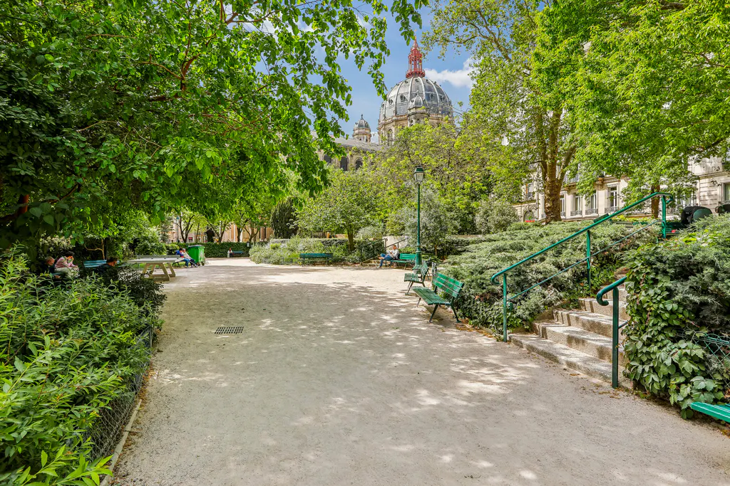 A park with green benches and trees, a gravel path, and a building with a dome in the background.