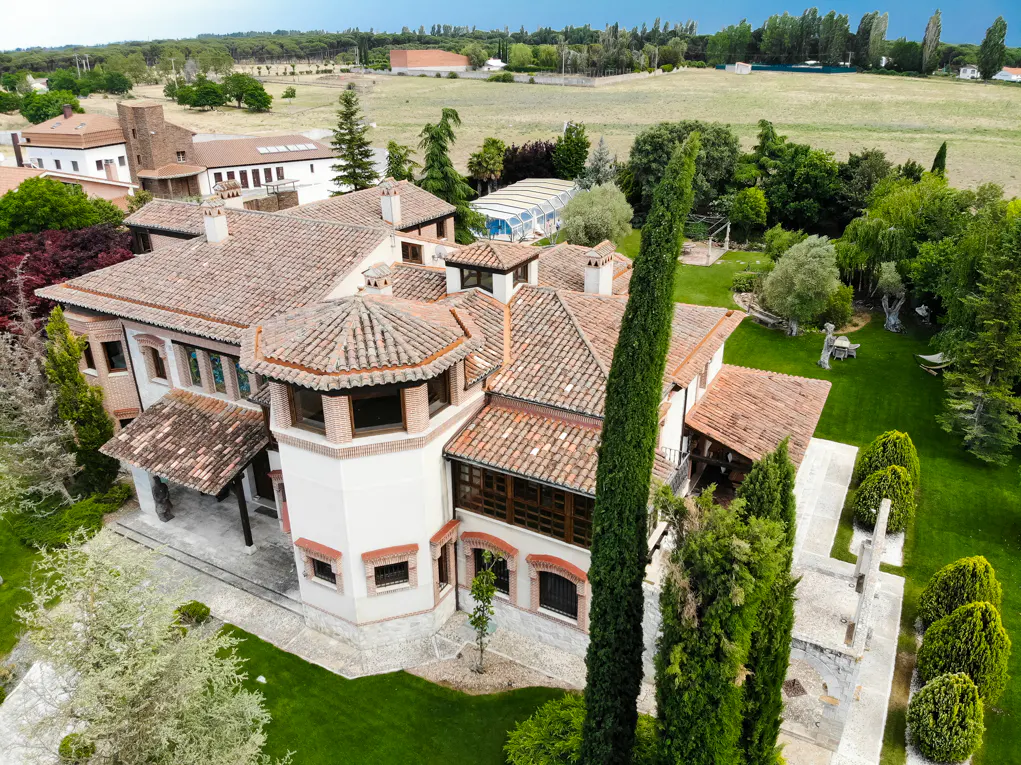 Aerial view of a large, multi-story villa with a terracotta tile roof, surrounded by lush green lawns and tall trees.
