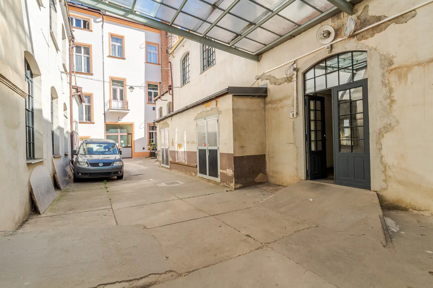 A paved courtyard between old buildings with a parked grey car, a glass canopy, and an open dark grey entrance door.