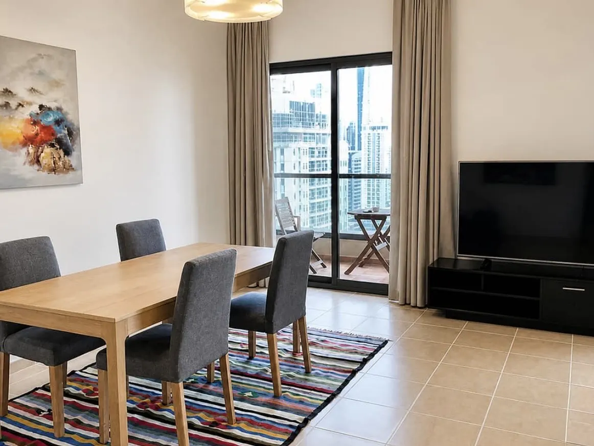 Apartment dining area with a light wood table, four gray chairs, and a colorful striped rug. Balcony with city view in the background.