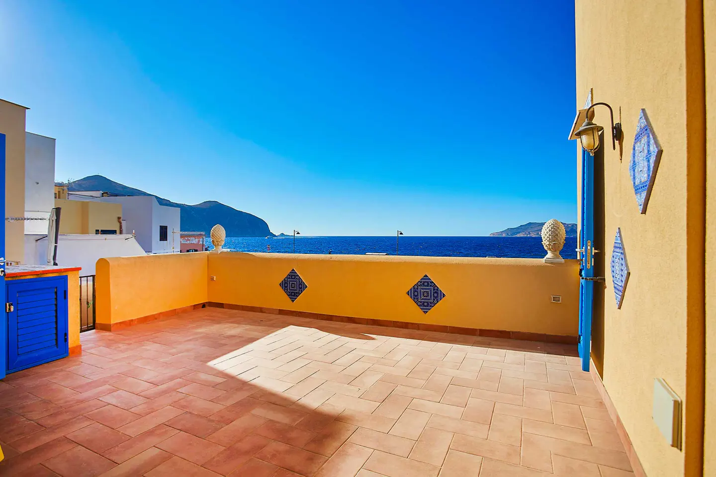 Terracotta-tiled patio with yellow walls, blue accents, and ocean view under a clear blue sky.