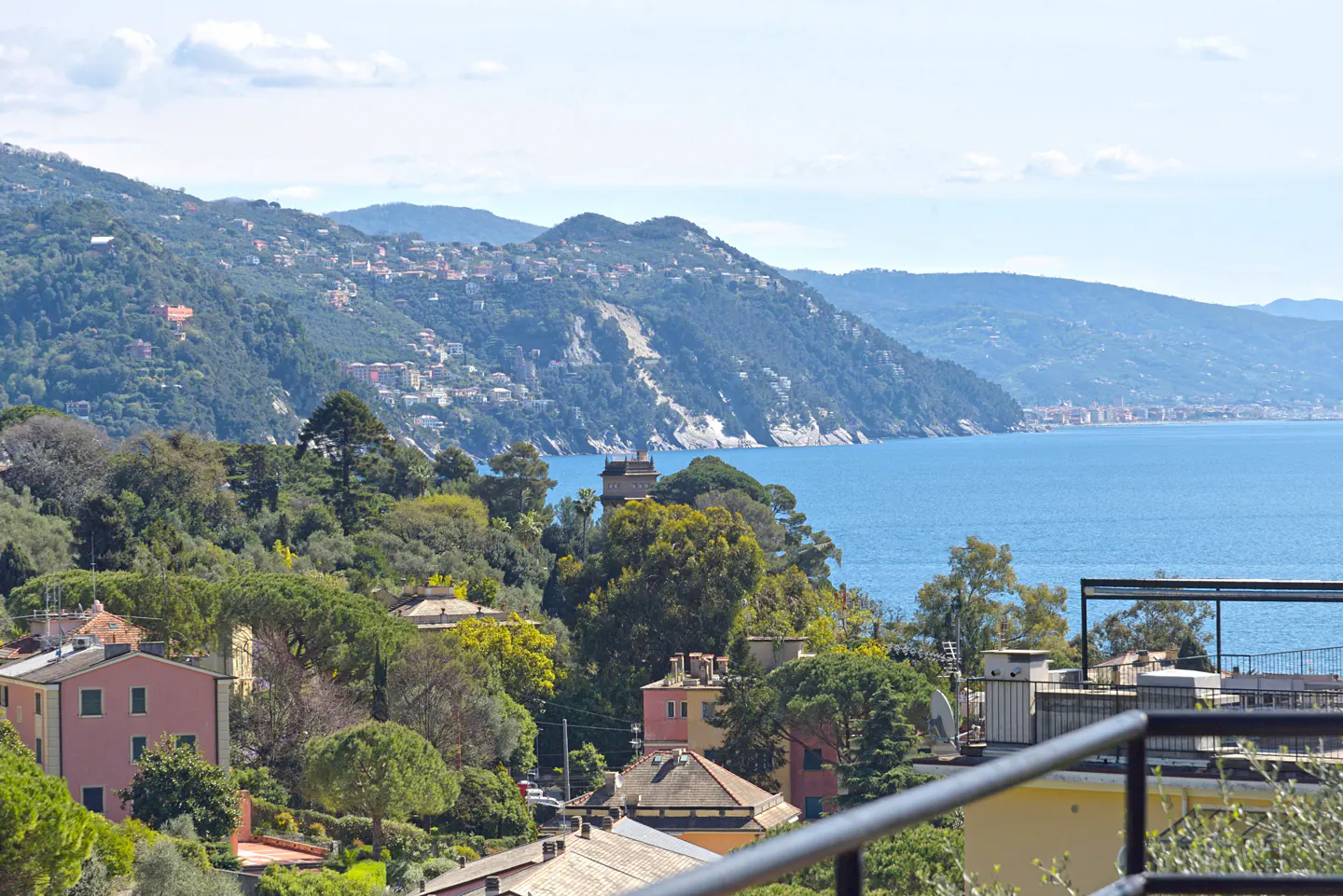 Scenic view of Italian coastline with mountains, blue sea, and colorful houses nestled among green trees. Balcony railing in foreground.