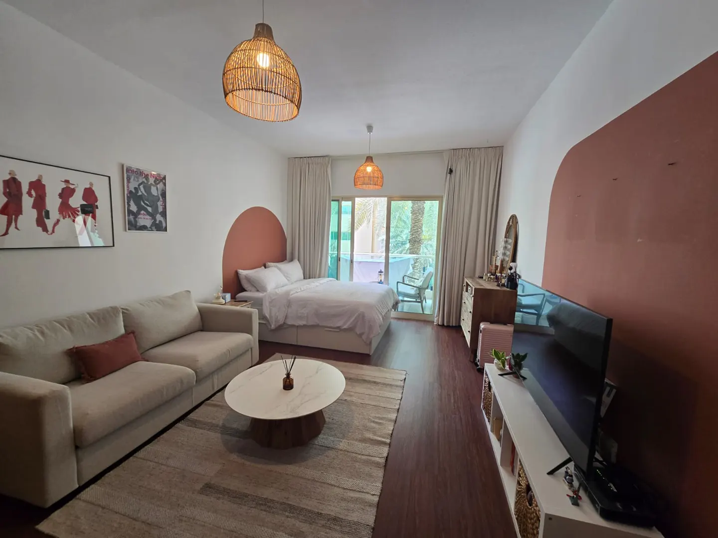 A bright studio apartment with a bed, sofa, TV, and balcony. The walls are white with a terracotta arch behind the bed. A woven pendant light hangs above.