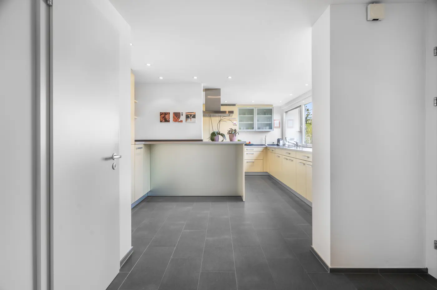 A bright kitchen with light wood cabinets and a gray tile floor is viewed through an open white door.