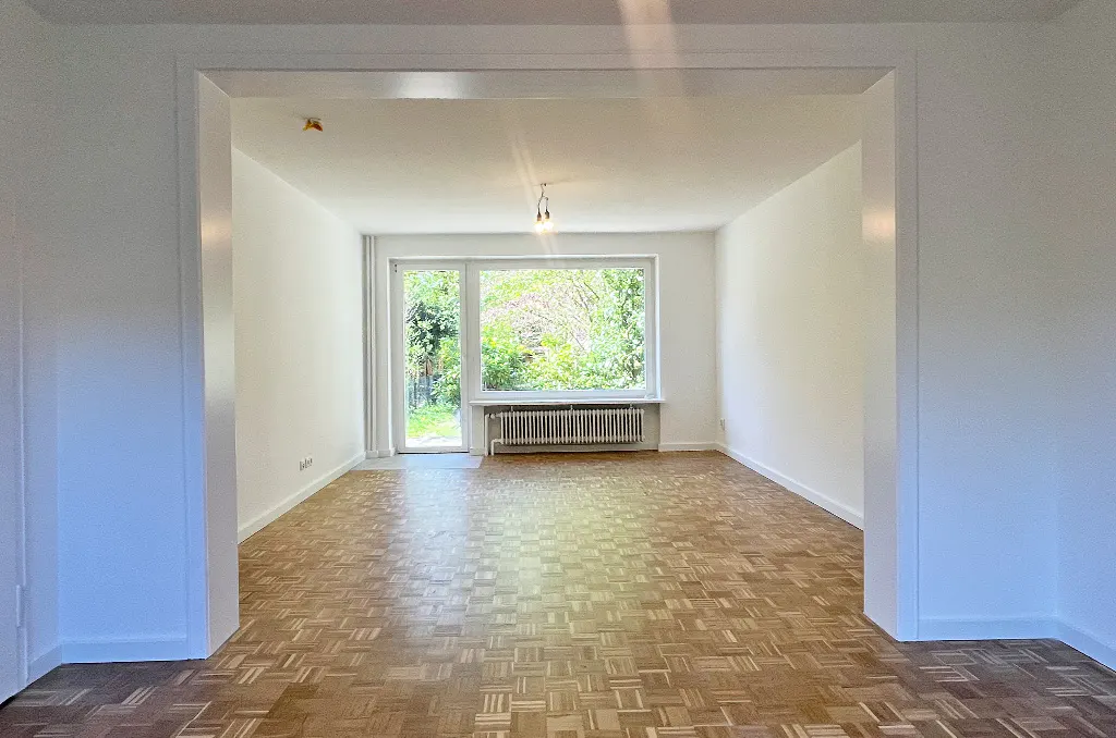 Empty room with parquet floor, white walls, and a large window overlooking a green garden. A radiator sits beneath the window.
