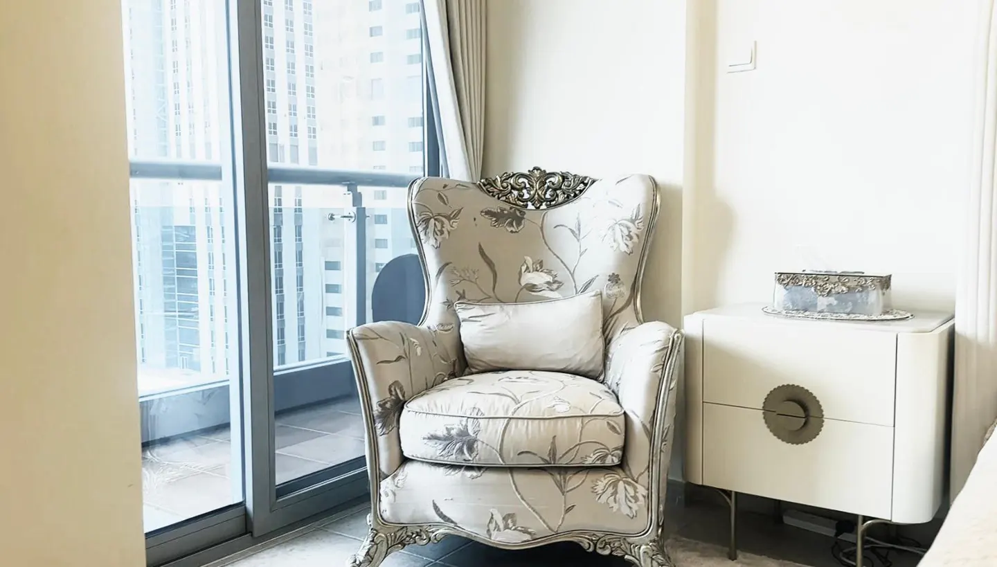 A floral armchair with a pillow sits near a window with a city view. A white nightstand with a decorative box is nearby.