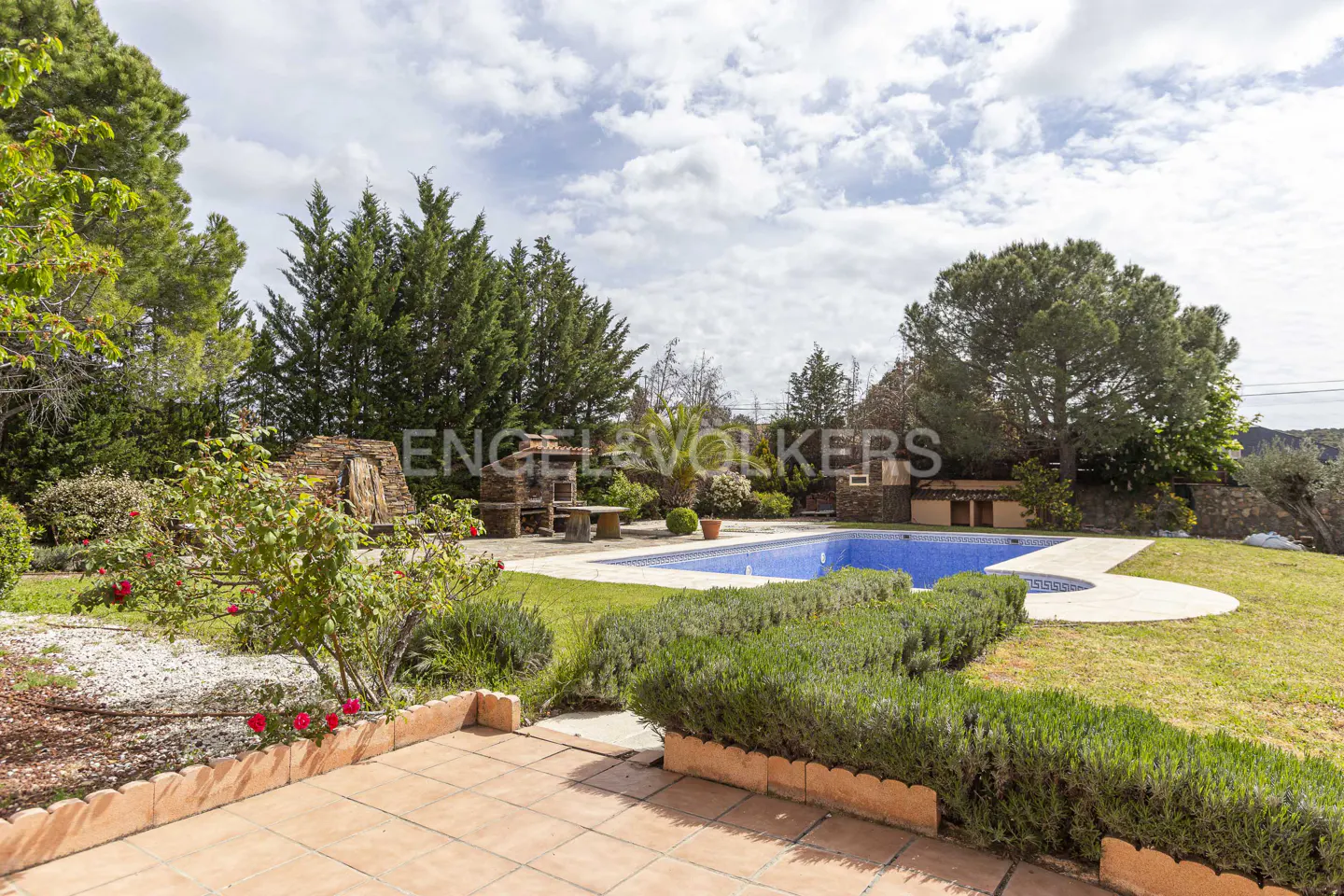 A backyard with a blue tiled pool, green lawn, stone BBQ, and trees under a cloudy sky.