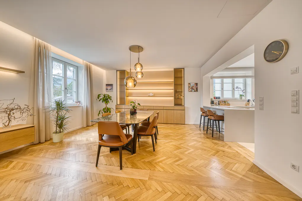 Bright, open dining room with herringbone wood floors, marble table, and tan leather chairs. Kitchen island visible in the background.
