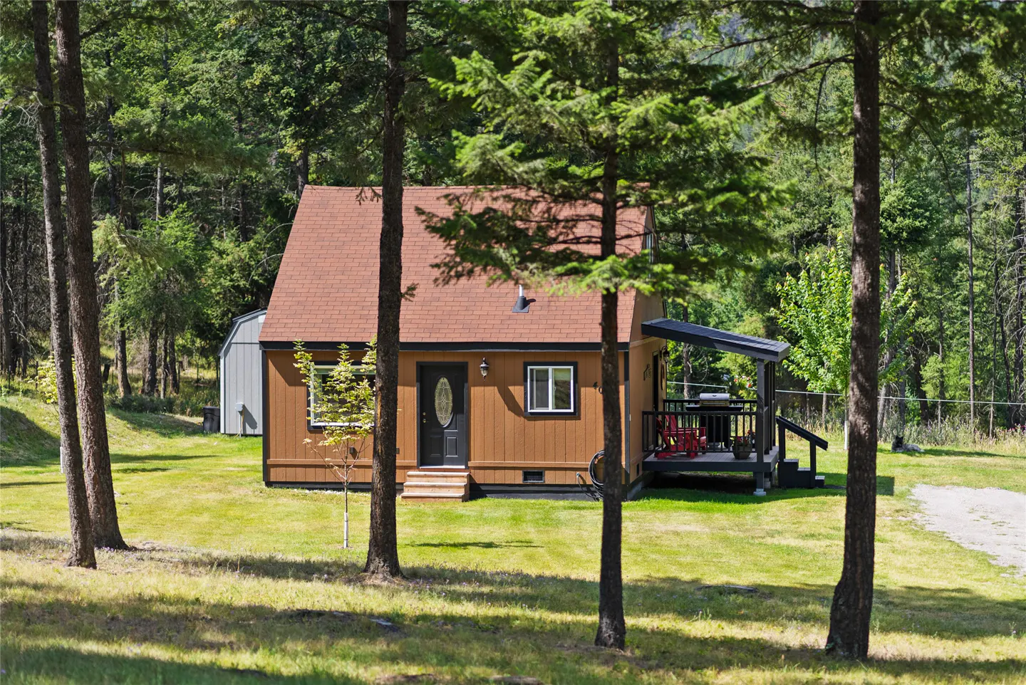 A brown house with a red roof sits on a green lawn, framed by tall trees and a forest backdrop.