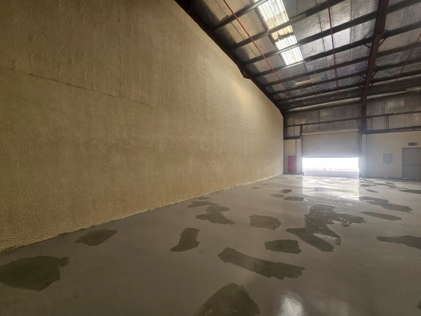 Empty warehouse with a beige wall, gray floor with water stains, and a partially open garage door.