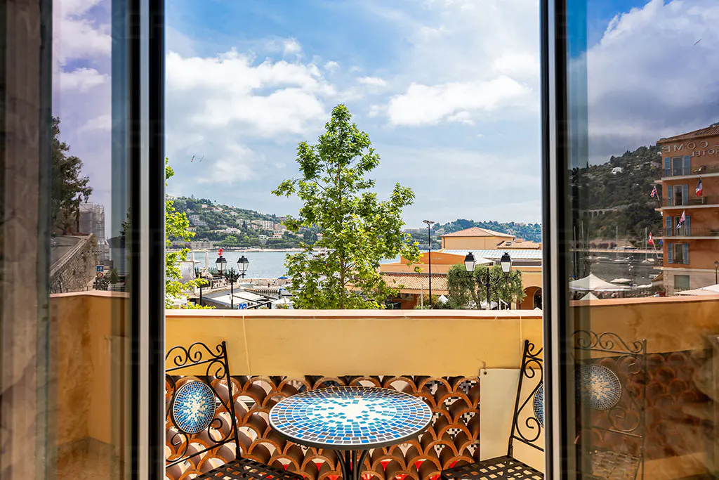 View from a balcony with a blue mosaic bistro table and chairs overlooking a harbor, trees, and coastal buildings.
