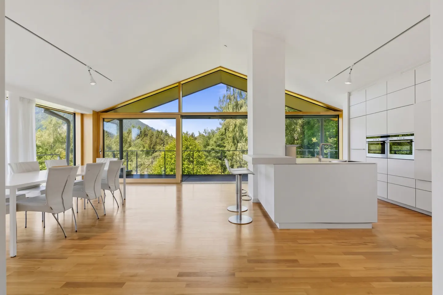 Modern open-plan kitchen and dining area with white cabinets, hardwood floors, and large windows overlooking green trees.