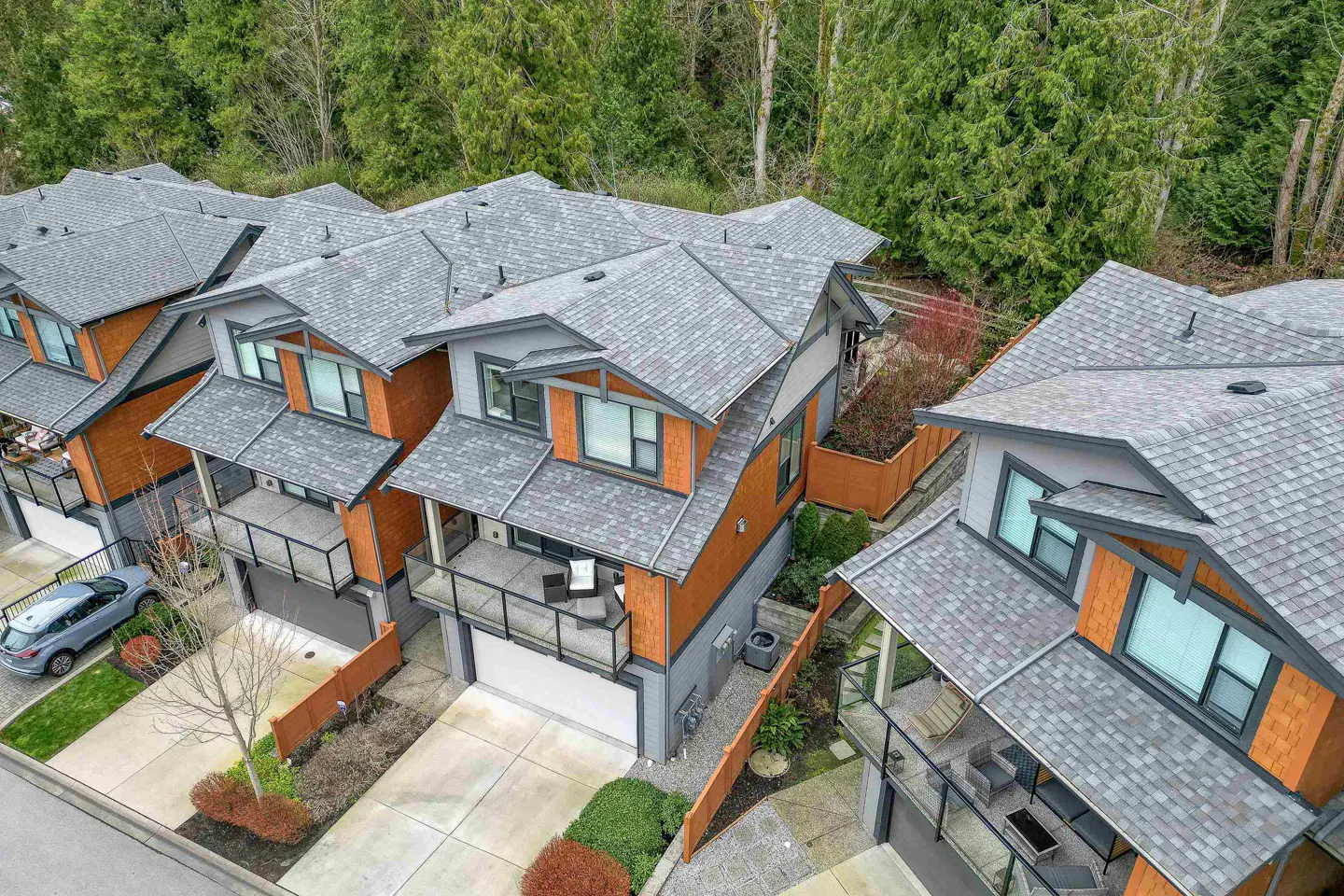 Aerial view of modern townhouses with gray roofs, wood and gray siding, balconies, and driveways, surrounded by green trees.