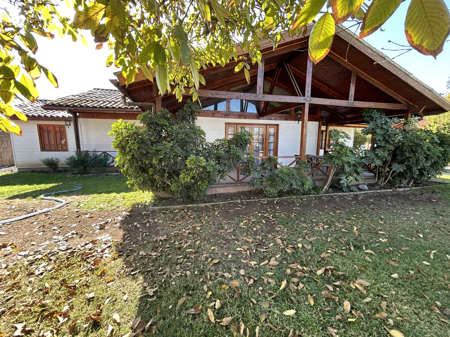 Exterior of a white house with a brown wood porch and roof. Green bushes line the front of the house. The lawn is green with fallen leaves.