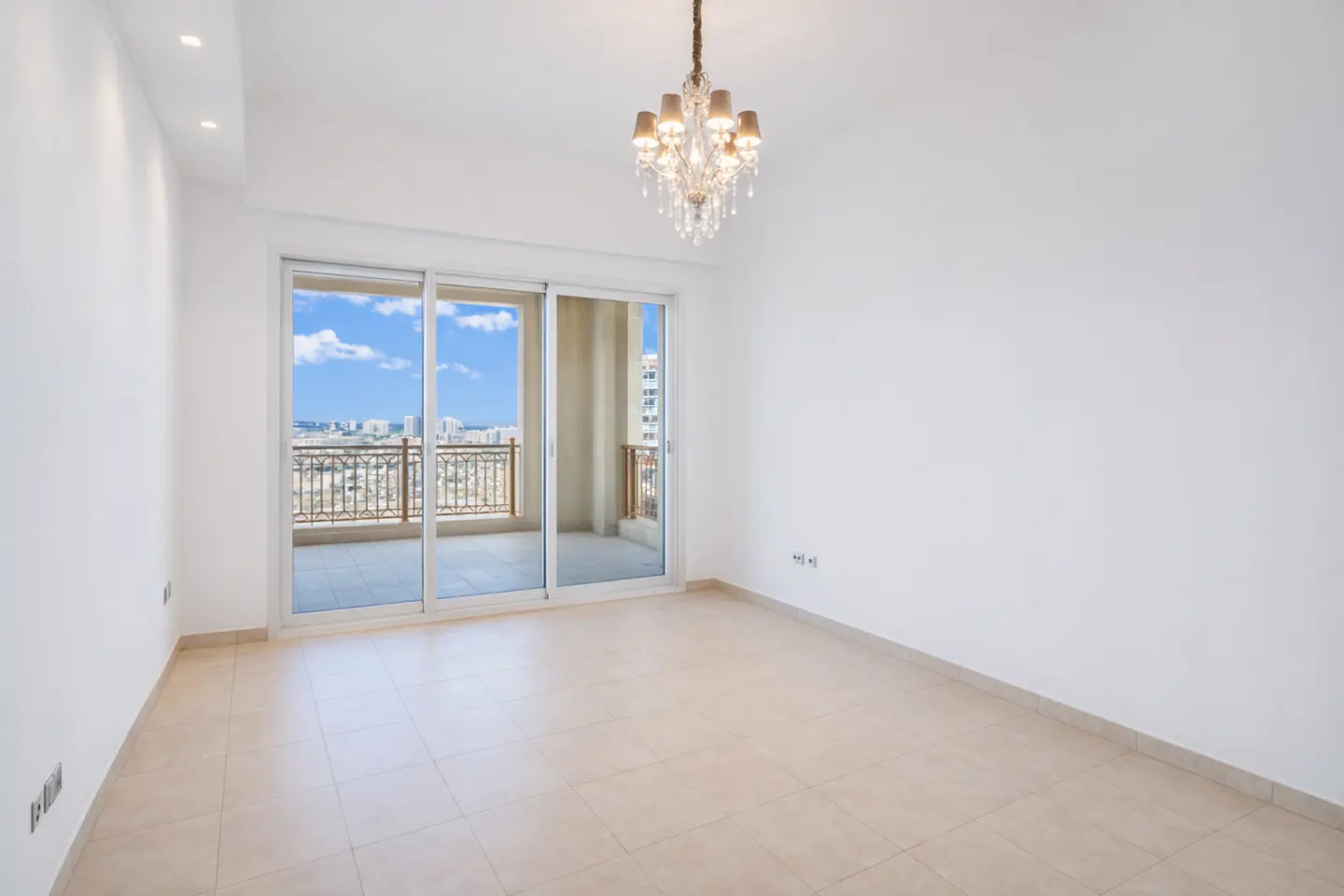 Bright, empty room with white walls, beige tile floor, and a chandelier. Sliding glass doors lead to a balcony with a city view.