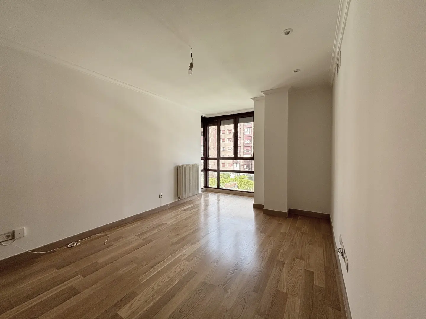Empty room with light wood floors, white walls, and a large window. A radiator sits under the window.