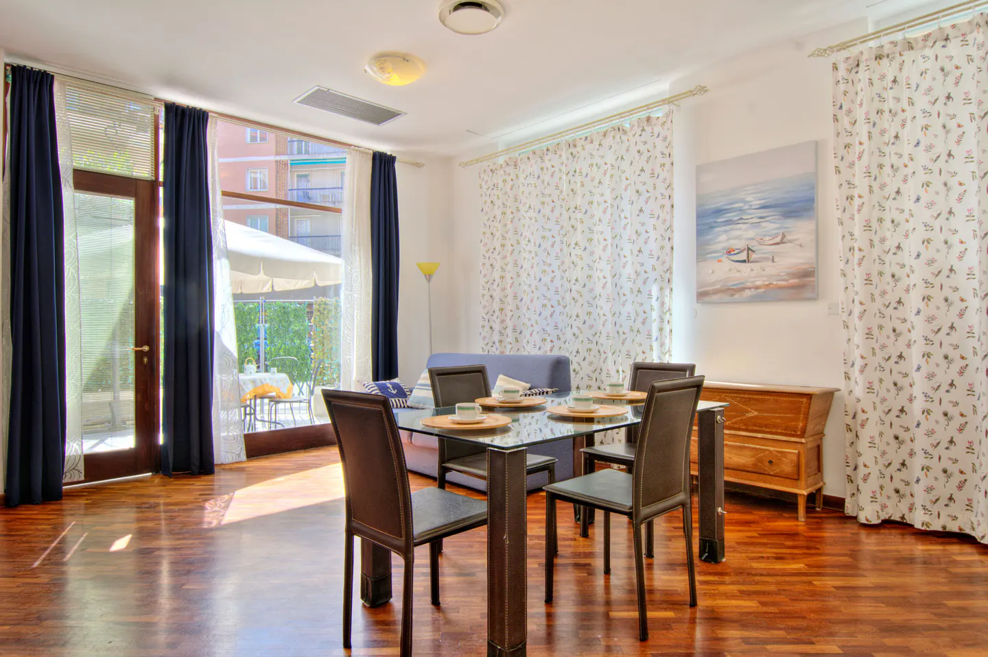 A dining room with a glass table, brown chairs, and hardwood floors. Blue curtains frame a door to a patio.