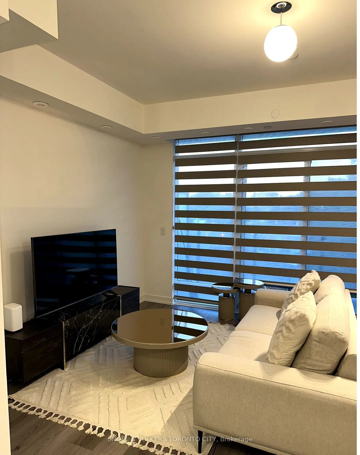 Living room with a white sofa, round coffee table, and a black TV stand with a large TV. The room has a large window with horizontal blinds.