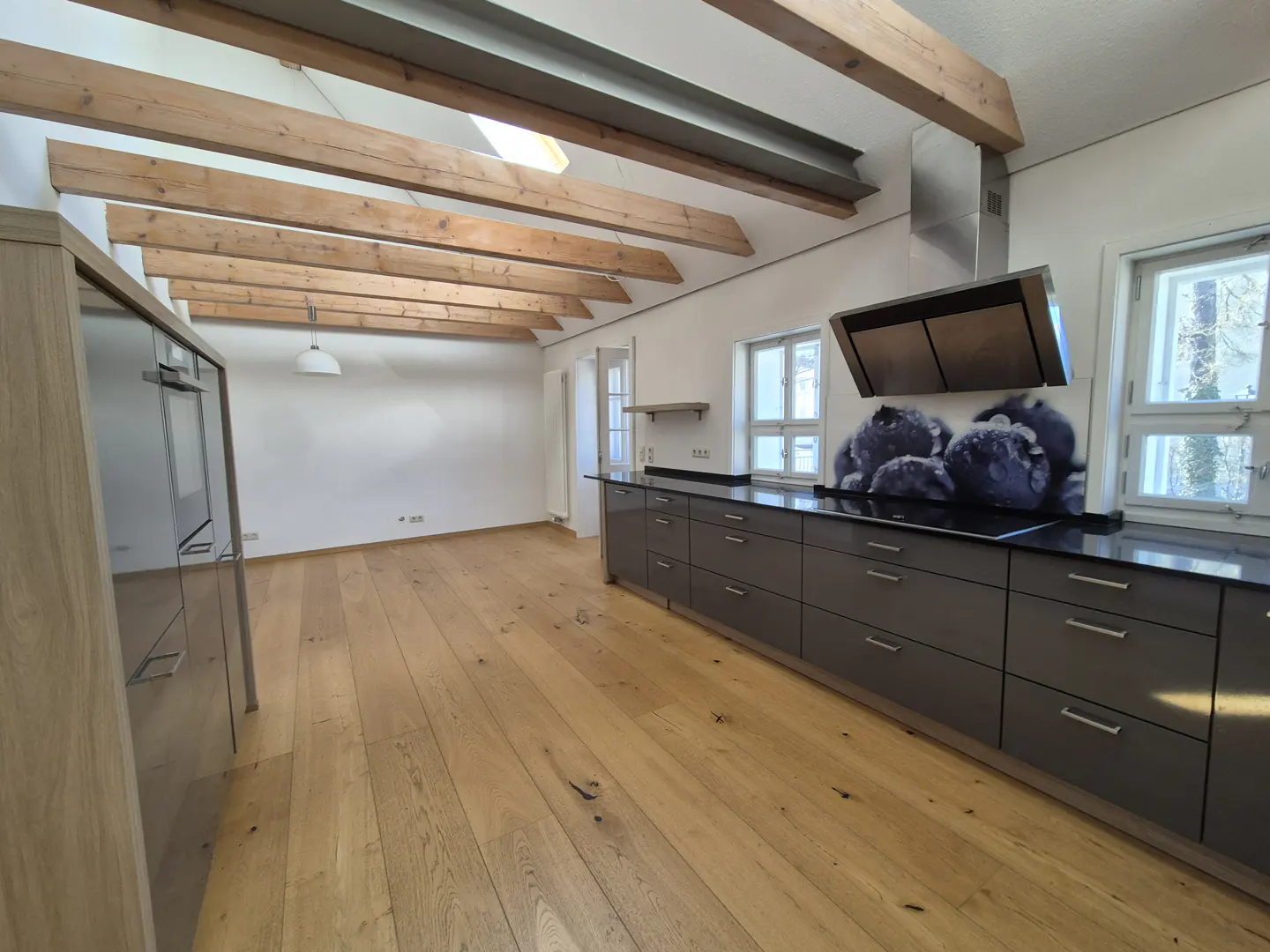 A bright, modern kitchen with wood floors and exposed beams. Gray cabinets line the wall, topped with a black countertop and blueberry backsplash.