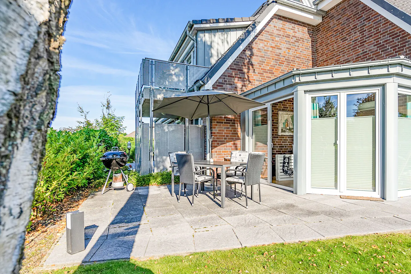 Outdoor patio with table, chairs, umbrella, and grill. Brick building with balcony and sliding glass doors in the background.