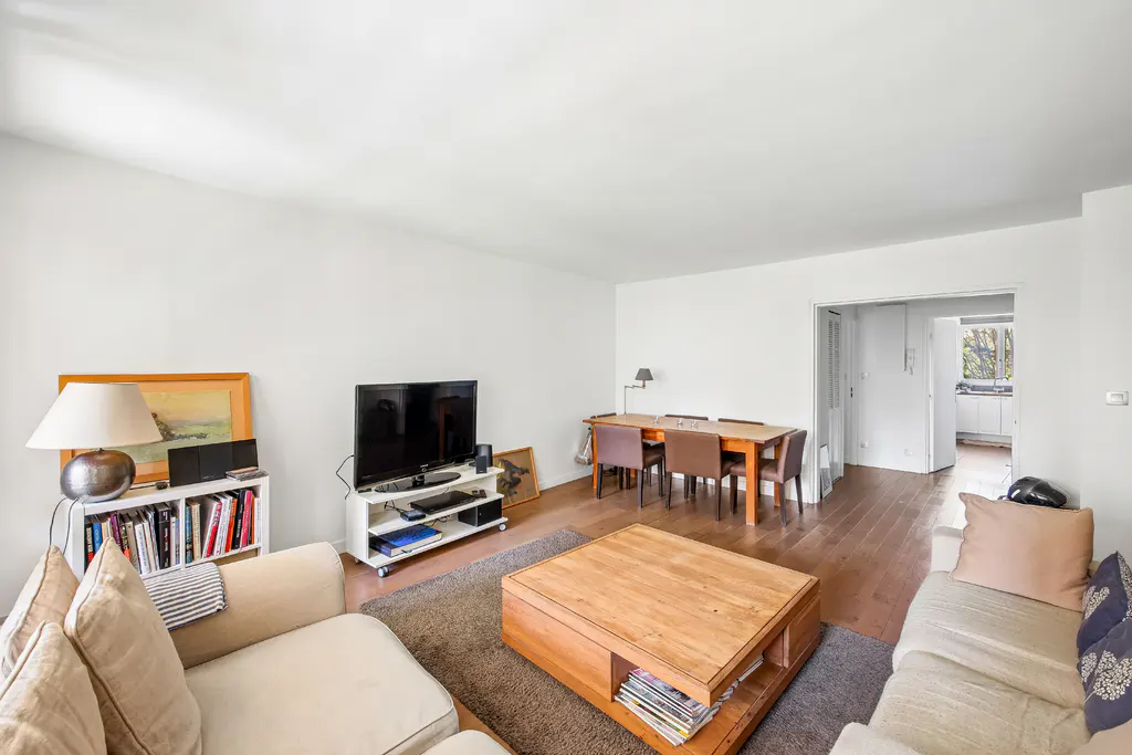 Bright living room with hardwood floors, white walls, and neutral furniture. A dining table and TV are visible.