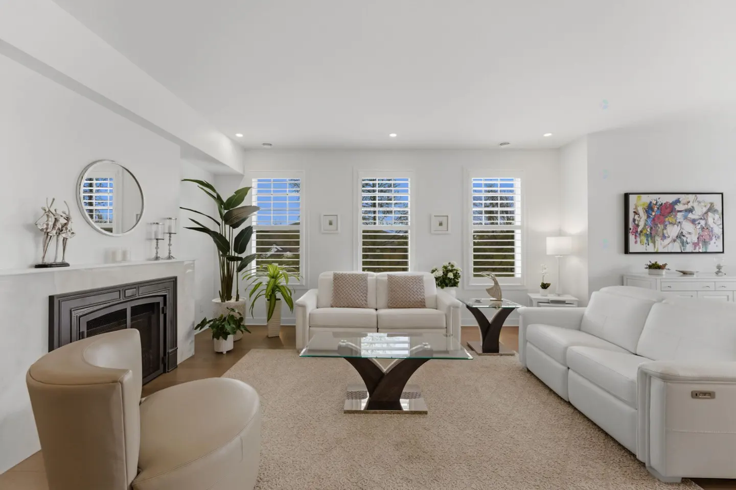 Bright living room with white sofas, a glass coffee table, and a fireplace with a round mirror above. Three windows with white shutters let in natural light.